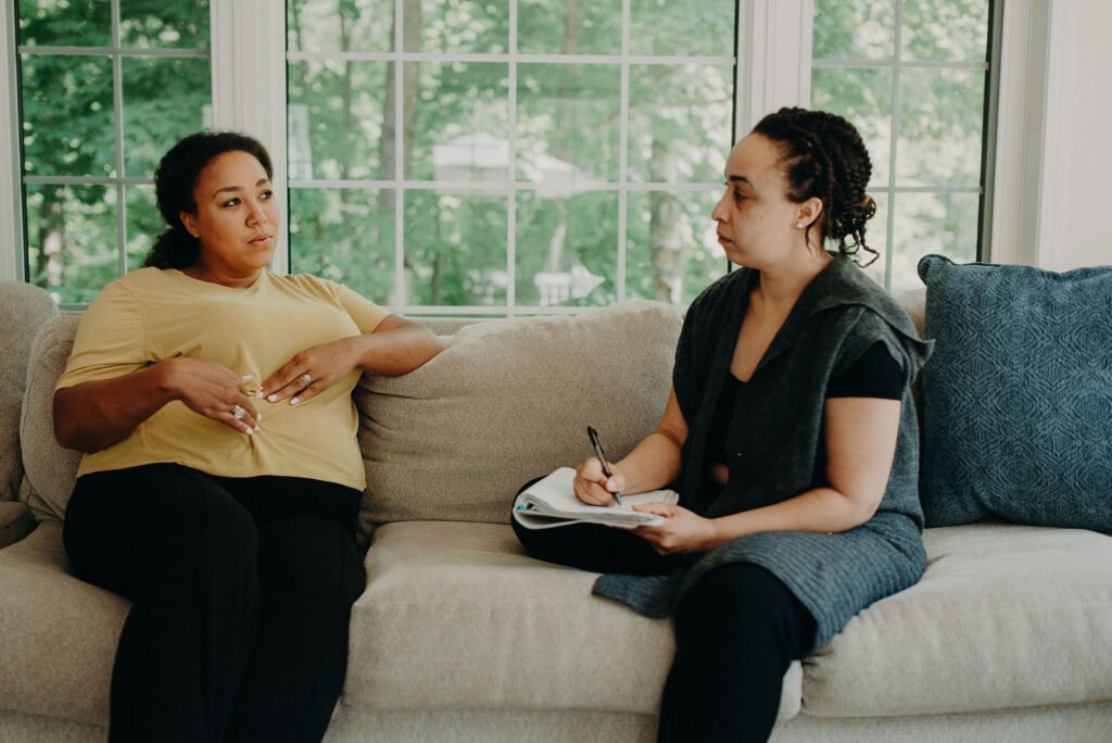 A pregnant woman in a yellow shirt talks to her doula in a green shirt on a beige couch.