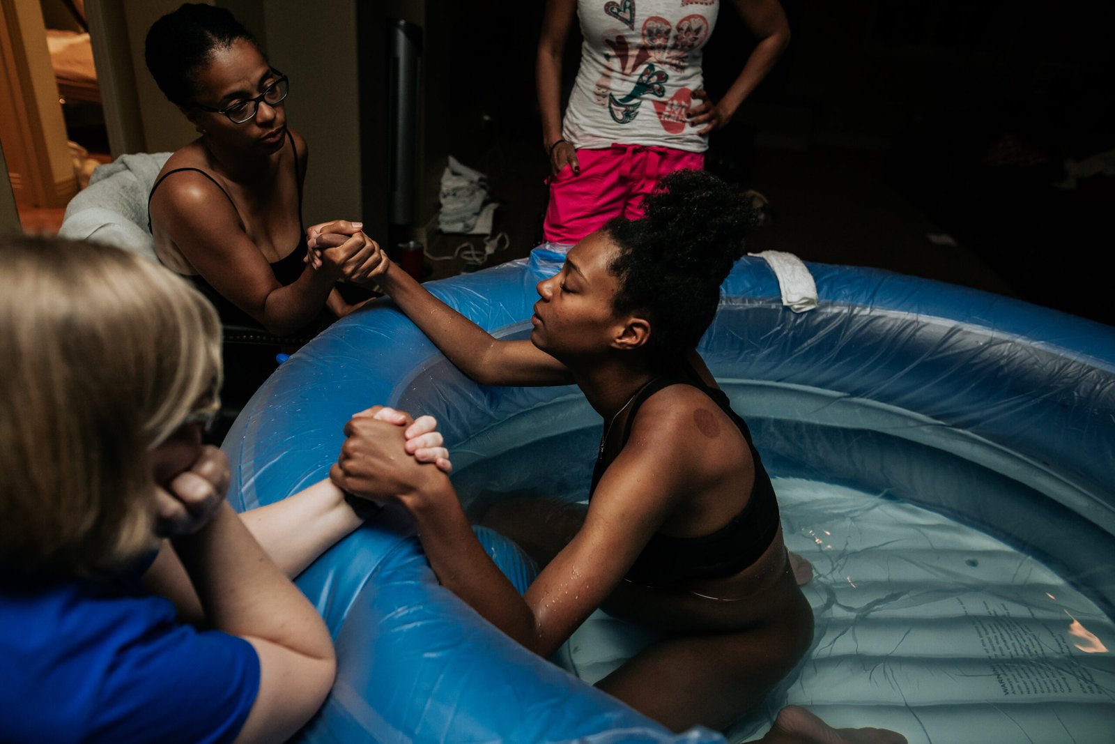 Birthing person in a blue tub with water holding hands with two doulas